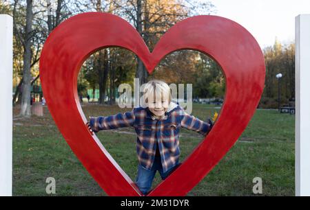Ein süßer, lächelnder Junge, 5-6 Jahre alt, in einem karierten Hemd in einem großen roten Herz, sieht glücklich in der Kamera aus. Muttertag, valentinstag Stockfoto
