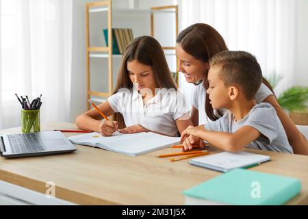 Online-Unterricht mit Laptop hilft mutter ihren Kindern dabei, Hausaufgaben am Schreibtisch zu machen. Stockfoto