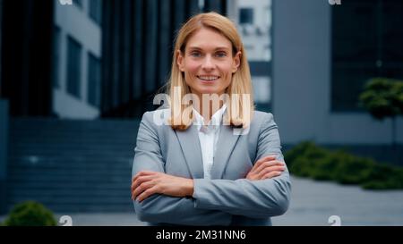 Selbstbewusste junge Geschäftsfrau im formellen Anzug, die sich mit gekreuzten Armen im Freien posiert und eine ehrgeizige unabhängige Anführerin lächelt Stockfoto
