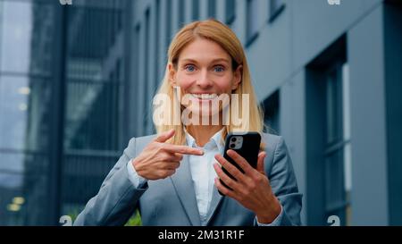 Kaukasische Geschäftsfrau hält Telefon steht im Freien enthusiastische glückliche Frau zeigt auf Smartphone-Bildschirm immer gute Nachrichten erhält profitabel Stockfoto