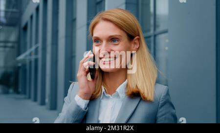 Fröhliche, enthusiastische, aufgeregte Frau, die im Freien mit ihrem Mobiltelefon telefoniert und ein gutes Erlebnis mit einem Freund teilt, der aus der Ferne über ein Smartphone chattet Stockfoto