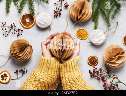 Handgefertigte Wabenkugel aus weißem und braunem Kaffeefilter in Frauenhänden. Skandinavisches Handwerkspapier Konzept. Draufsicht. Stockfoto