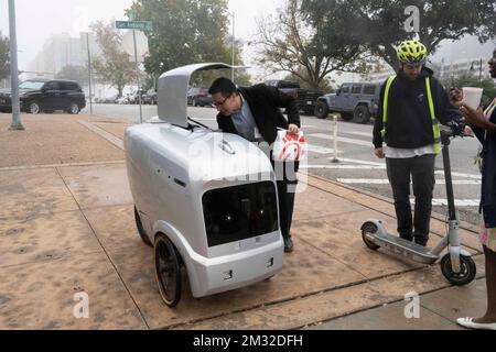 Ein autonome Lieferfahrzeug, hergestellt von der in Austin ansässigen Refraktion AI, liefert ein Chick-fil-A Mittagessen an die Mitarbeiter des Gerichtsgebäudes von Travis County unter der Aufsicht eines Testpiloten, der dem Fahrzeug auf einem Roller folgt. Die selbstfahrenden Roboter werden seit Monaten auf den Straßen von Austin getestet. Stockfoto