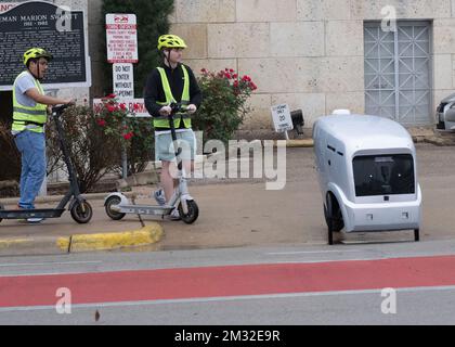 Ein autonome Lieferfahrzeug, hergestellt von der in Austin ansässigen Refraktion AI, liefert ein Chick-fil-A Mittagessen an die Mitarbeiter des Gerichtsgebäudes von Travis County unter der Aufsicht eines Testpiloten, der dem Fahrzeug auf einem Roller folgt. Die selbstfahrenden Roboter werden seit Monaten auf den Straßen von Austin getestet. Stockfoto