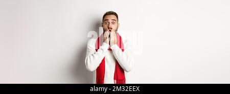 Weihnachtsferien und Feier Konzept. Schöner bärtiger Kerl in rotem Schal und Pullover, der schockiert, beeindruckt von Werbung, weiß Stockfoto