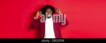 Weihnachtseinkäufe und People-Konzept. Happy Black Mann feiert neues Jahr, trägt Party-Outfit, berühren Hut und lächeln, stehen gegen rot Stockfoto