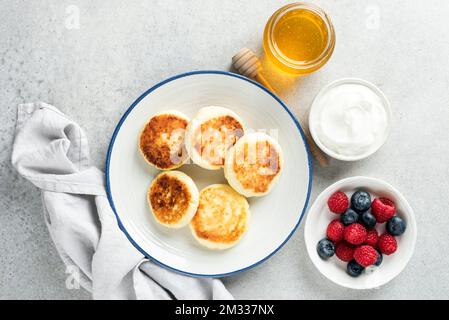 Käsepfannkuchen, Syrniki serviert mit Beeren, Sauerrahm und Honig. Süße Frühstückskäse Pfannkuchen in Öl gebraten. Draufsicht Stockfoto