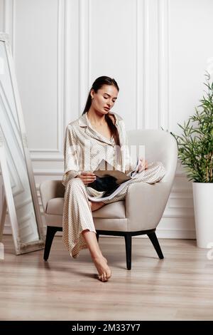 Frau im Pyjama sitzt im Stuhl und liest Zeitschrift Stockfoto