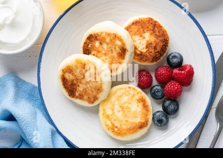 Süße Frühstückskäse, Pfannkuchen oder Fritters, Syrniki serviert mit Beeren auf weißem Teller, Blick von oben Stockfoto