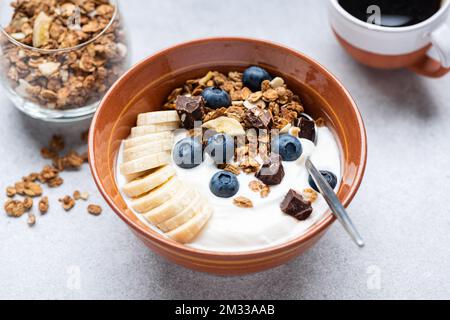 Joghurt mit Granola, Banane, Blaubeeren und dunkler Schokolade in der Schüssel, grauer Kopfsalat-Tisch, Nahaufnahme. Gesundes Essen Stockfoto