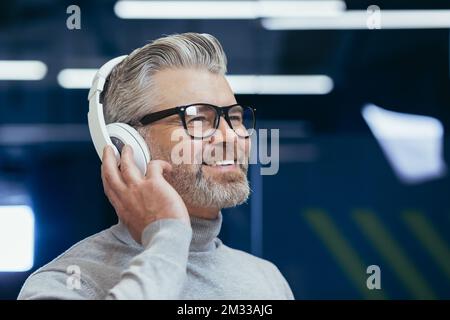Nahaufnahme. Porträt eines älteren grauhaarigen Mannes mit Brille, der im Büro Musik über Kopfhörer hört, Kopfhörer in der Hand hält, lächelt, wegschaut. Stockfoto