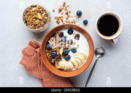 Joghurt mit Müsli, Banane, Blaubeeren und dunklen Schokoladenstückchen in einer Schüssel serviert mit einer Tasse schwarzen Kaffees. Top View gesundes Frühstück Stockfoto