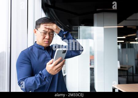 Aufgewühlter asiatischer Mann im Büro, der schlechte Nachrichten aus dem Telefon im Internet liest, Geschäftsmann im Hemd am Fenster, der im Büro arbeitet, mit Smartphone. Stockfoto