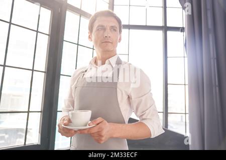 Ein männlicher Kellner in einer grauen Schürze hält eine Tasse Kaffee. Der Barista serviert eine Tasse heißen Kaffee vor dem Hintergrund eines großen Fensters mit Platz zum Kopieren. Hochwertiges Foto Stockfoto