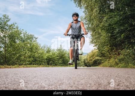 Porträt eines glücklichen lächelnden Mannes in Radbekleidung, Helm und Sonnenbrille, der auf dem asphaltierten Radweg außerhalb der Stadt ein Fahrrad fährt. Aktive spo Stockfoto