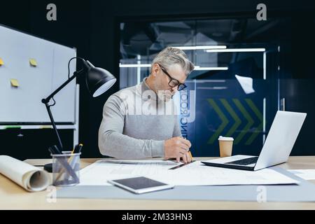 Ein gut aussehender, grauhaariger Senior Architekt, Designer, Ingenieur macht eine Zeichnung mit einem Bleistift auf Papier, macht einen Plan, konzentriert sich auf das Projekt am Tisch im Büro. Stockfoto