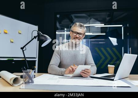 Ein älterer, grauhaariger Mann, Architekt, Designer, Ingenieur. Im Büro mit einem Laptop und Papierkram sitzen. Er hält ein Notizbuch mit einem Bleistift in der Hand. Er sieht in die Kamera und lächelt. Stockfoto