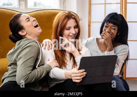Fröhliche, multikulturelle weibliche Freunde, die sich zu Hause in ihrer Freizeit aufhalten – Fröhliche junge Frauen, die sich im Wohnzimmer Videos oder tv-Serien auf einem Tablet ansehen Stockfoto