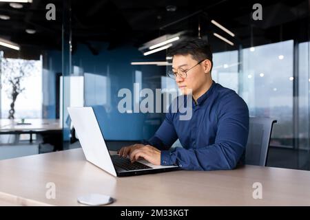Seriöser und umsichtiger Geschäftsmann, der im Büro arbeitet, am Tisch sitzt und ein Notebook benutzt, ein erfahrener asiatischer Chef im Hemd denkt und auf der Tastatur tippt. Stockfoto