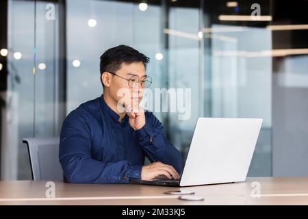 Seriöser und umsichtiger Geschäftsmann, der im Büro arbeitet, am Tisch sitzt und ein Notebook benutzt, ein erfahrener asiatischer Chef im Hemd denkt und auf der Tastatur tippt. Stockfoto