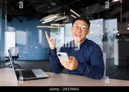 Porträt eines glücklichen asiatischen Geschäftsmanns, Mann, der Erfolg und Sieg feiert, Boss schaut in die Kamera im Büro, hält Telefon, Unternehmer, der im Büro arbeitet. Stockfoto