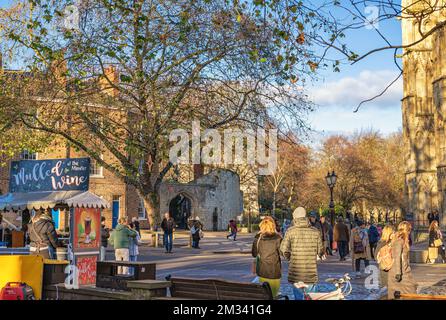 Touristen stehen um einen offenen Bereich neben alten Gebäuden und in der Nähe eines übergeordneten Baumes. Ein Glühwein-Kiosk befindet sich im Vordergrund. Stockfoto