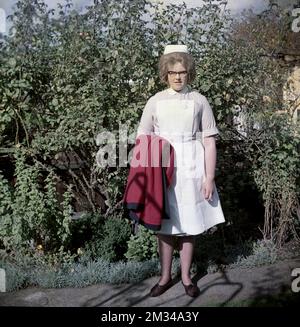 1960er, historisch, steht draußen für ihr Foto, eine junge Krankenschwester in ihrer Uniform, ein kurzärmeliges, gestreiftes Fliederkleid mit Kragen und weißer Schürze, hält ihren Schwesternumhang mit kastanienbraunem Futter, England, Großbritannien. Auf ihrem Kopf befindet sich eine traditionelle Schwesternmütze, und sie trägt die Brille, die in den 50s und 60s Jahren modisch war, auch als Katzenaugenbrille bekannt. Stockfoto