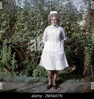 1960er, historisch, steht draußen für ihr Foto, eine junge weibliche Krankenschwester in ihrer Uniform, ein kurzärmeliges, gestreiftes Fliederkleid, mit Kragen und weißer Schürze, mit einer Schwesternuhr, England, Großbritannien. Auf ihrem Kopf befindet sich eine traditionelle Schwesternmütze, und sie trägt die Brille, die in den 50s und 60s Jahren modisch war, auch als Katzenaugenbrille bekannt. Stockfoto