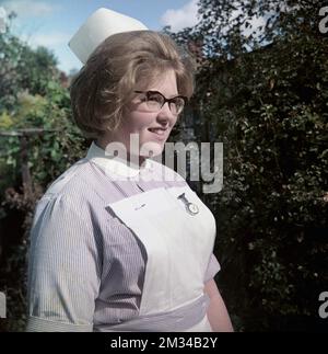 1960s, historical, a young female nurse standing outside in her uniform, a  short sleeve striped lilac dress, with collar and white apron, with a nurses fob watch attached, England, UK. On her head is a traditional nurse cap and on her face she is wearing the spectacles that were fashionable in the 50s and 60s, known as Cat Eye glasses. Stock Photo