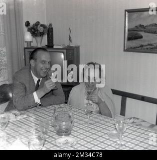 1960er Jahre, historisch, ein Mann und eine Dame sitzen an einem Tisch in einem Vorderzimmer und genießen ein Glas Wein, England, Großbritannien. In der Ecke ist ein Fernsehgerät aus dieser Zeit zu sehen. Stockfoto