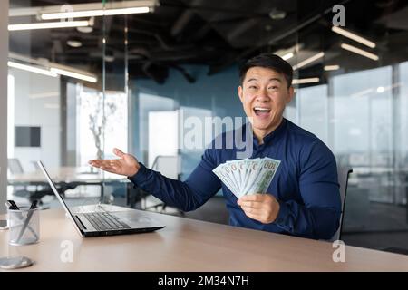 Glücklicher asiatischer Banker, der in einem modernen Bürogebäude arbeitet, ein Mann, der in die Kamera schaut und glücklich ist, Geld in bar zu zeigen, Dollar Deal-Einnahmen. Stockfoto