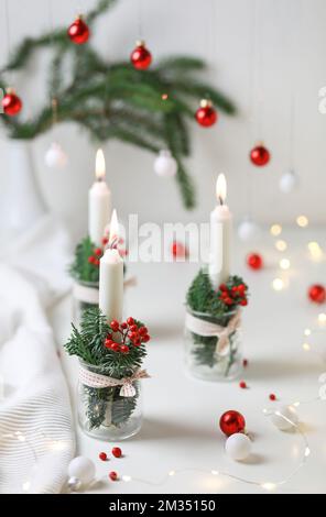 Weihnachtsdekoration. Kerzen in Glasgefäßen, dekoriert mit Tanne und roten Beeren auf einem weißen Tisch. Stockfoto