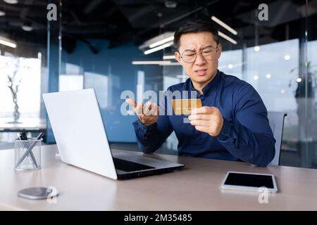 Betrogener Geschäftsmann, enttäuschter Mann, der im Büro saß, asiatischer Mann, der versucht, Banküberweisungen zu tätigen und im Online-Shop einzukaufen, nicht genug Geld mit Bankkreditkarte. Stockfoto