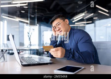 Betrogener Geschäftsmann, enttäuschter Mann, der im Büro saß, asiatischer Mann, der versucht, Banküberweisungen zu tätigen und im Online-Shop einzukaufen, nicht genug Geld mit Bankkreditkarte. Stockfoto