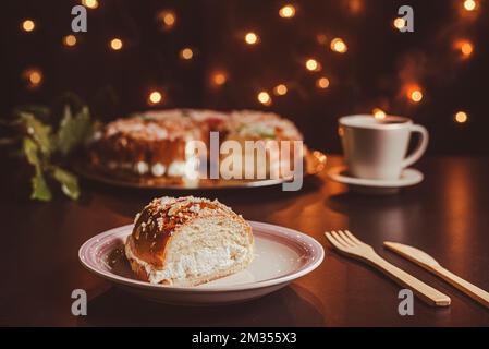 Ein Stück roscón de reyes auf einem weißen und rosafarbenen Teller auf einem dunklen Tisch mit Holzbesteck, im Hintergrund ein roscón de reyes und eine Tasse Kaffee Stockfoto