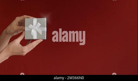 Eine Frauenhand hält eine Geschenkbox auf einem rot-burgunderfarbenen Hintergrund. Minimalismus. Speicherplatz kopieren. Zeit für Geschenke Stockfoto
