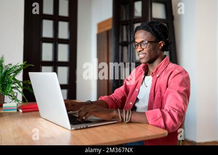 Glücklicher, zufriedener junger afrikanischer Freiberufler, der im Heimbüro sitzt und die Arbeit aus der Ferne genießt Stockfoto