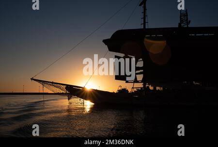 Ein altes Fischerboot liegt am Hafen vor einem großen Metallschiff, gegen das Licht. Die Sonne geht unter. Der Himmel ist blau und rot. Stockfoto