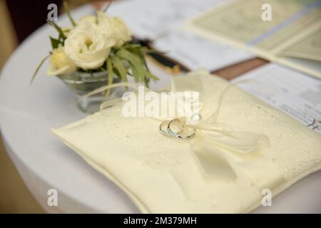 Zwei Platinringe für die Hochzeit liegen auf einem Seidenspitzenkissen für Ringe. Hochzeitszubehör Braut und Bräutigam, bevor die Zeremonie auf einem weißen Tisch fotografiert wurde Stockfoto