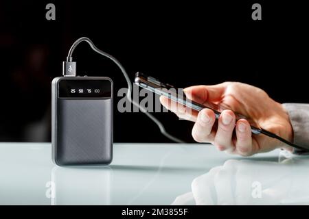 Schwarze Powerbank mit schwarzem Kabel zum Aufladen eines Smartphones in männlichen Händen auf einem schwarzen. Zusätzliche Stromversorgung. Minimalistisches Bild Stockfoto