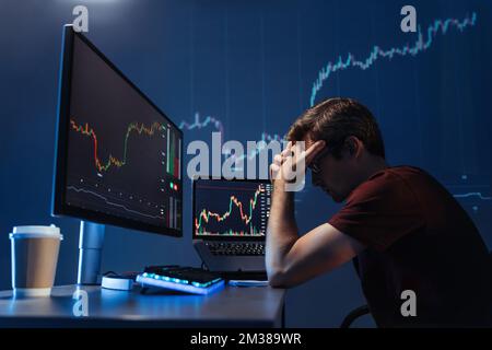 Seitenansicht eines deprimierten, rücksichtsvollen männlichen Krypto-Investors, der Kopf in den Händen vor dem Computer hält, mit einem Kerzenständerdiagramm des Krypto-Währungsmarktes, gescheitert, Geld zu gewinnen, aufgebracht durch die globale Rezession Stockfoto
