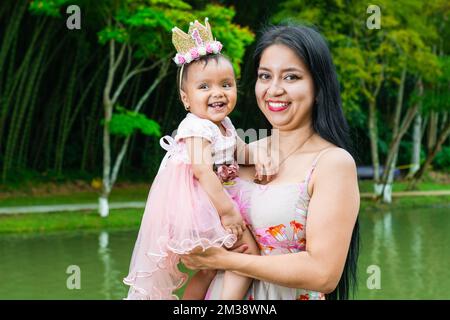 Porträt einer Mutter, die ihr Baby in den Armen hält, während sie glücklich auf einen See und einen riesigen Bambuswald im Hintergrund lächelt. Stockfoto