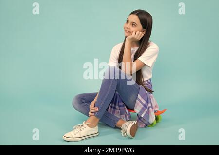 Fröhlicher, modiger Teenager. Modische selbstbewusste Teenager-Mädchen tragen trendige T-Shirt und Jeans sitzen auf Skateboard, posiert auf blauem Hintergrund. Mode Stockfoto