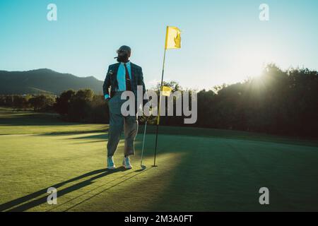 Ein Mann mit Glatze und Bart, mit altem Geld, trägt eine Sonnenbrille, raucht eine Zigarre und posiert mit einem Golfschläger in der Hand, neben einem Fahnenstab an einem sonnigen Vater Stockfoto