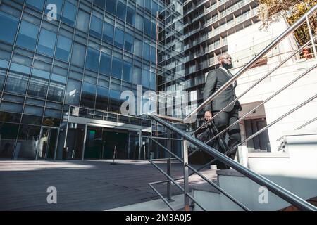 Ein Foto eines afrikanischen Arbeiters, der die Treppe mit einem Metallgeländer hochklettern will, während er eine Wochenendtasche trägt, eine Brille trägt und eine dunkle trägt Stockfoto