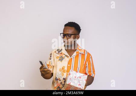 Ein junger afroamerikanischer Mann, der eine Brille trägt und sein Handy benutzt, mit einer Geschenkbox in der Hand, mit einem ernsthaften Look auf weißem Hintergrund Stockfoto