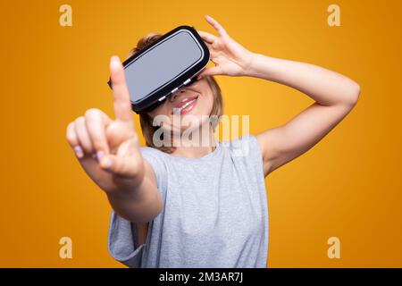 Smiling woman using finger to touch on imaginary panel viewing on VR device, isolated over yellow background. Metaverse technology concept. Stock Photo