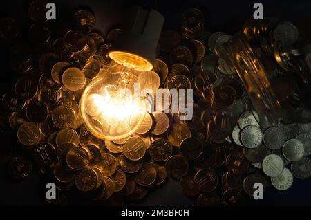 Das Konzept des Stromverbrauchs in diesen Krisenzeiten, dargestellt durch eine Glühbirne und verstreute Euro-Münzen, dunkler Hintergrund Stockfoto