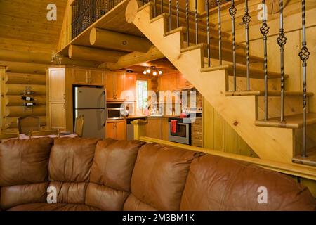 Wohnzimmer mit dunkelbraunem Ledersofa, Küche und Kiefernholztreppe in der Blockhütte. Stockfoto