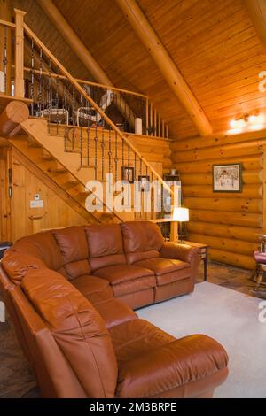 Dunkles braunes Ledersofa im Wohnzimmer und Treppe führt zum Hauptschlafzimmer oben in der Blockhütte. Stockfoto